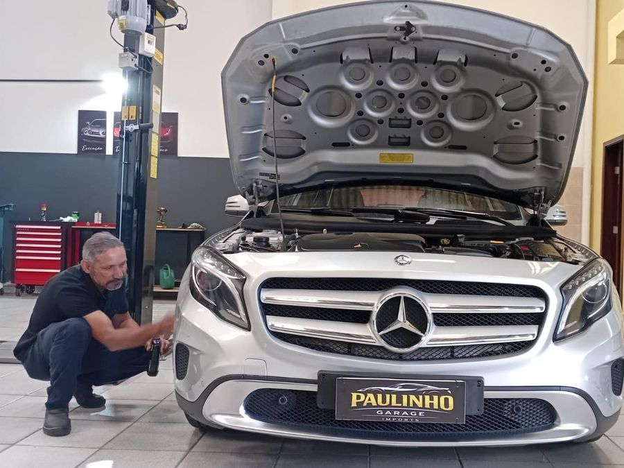 Paulo agachado atendendo uma Mercedes-Benz GLA prata com o capô aberto, no salão da oficina